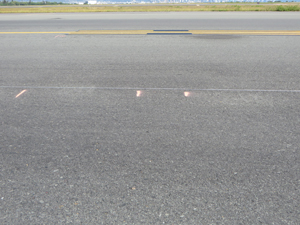 Overview photo (shot in the transverse direction) showing two corrugations. Orange transverse point lines on the pavement have been used to mark the start and stop locations of the corrugations. The corrugation area in between the paint marks is lighter in color due to water ponding.