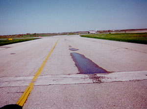 Overview photo of a taxiway pavement with visible low-severity rutting in one wheel path.            The rutting is visible because there is standing water in the wheel path.