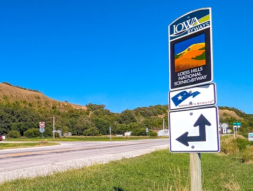 A highway lined with green grass and trees with an Iowa Byways sign