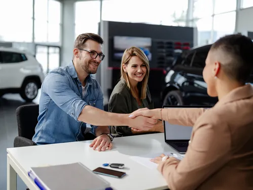 A car salesperson shakes hands with a male customer over a desk in an auto dealership