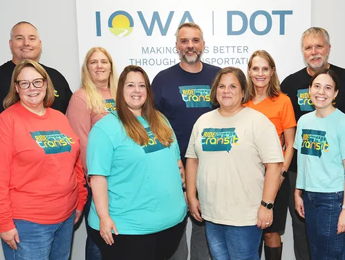 Group photo of nine Iowa DOT Transit staff members standing in front of an Iowa DOT sign and wearing colorful 'Ride Iowa Transit' shirts.