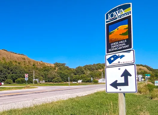 A highway lined with green grass and trees with an Iowa Byways sign