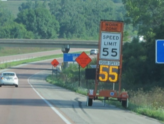 Speed feedback trailer displaying a speed limit sign of 55 and a digital speed readout along a roadside work zone.
